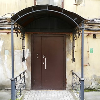 На фото: вход в здание, полуторная металлическая входная дверь с домофоном, над входом установлен защитный козырек на двух металлических столбах, перед входом сделана отмостка тротуарной плиткой, фасад здания оштукатурен